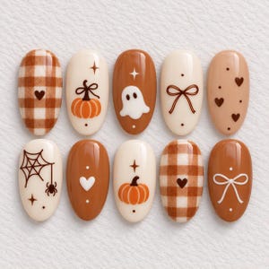 May include: Ten oval nail art tips featuring a seasonal theme. Designs include brown and white gingham patterns, orange pumpkins, a white ghost, brown bows, small hearts, a spider web with a spider, and star accents on cream and brown backgrounds.