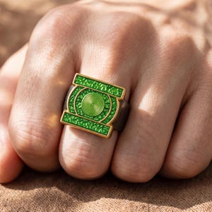 May include: A square-shaped ring featuring a bright green textured inlay and a central circular emblem with concentric rings. The ring has a gold-toned metal border and a dark, matte-finished band, showcasing a bold, geometric design.