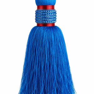 Puede incluir: Una borla decorativa con una gran cabeza esférica de color azul vibrante y largos flecos azules fluidos. El cuello está acentuado con una banda roja brillante y una sección central adornada con filas de pequeños cristales azules brillantes.