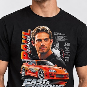 Op de afbeelding: Een zwart t-shirt met een grafische print van Paul Walker en een feloranje Toyota Supra. Het ontwerp bevat de tekst "Paul Walker", "Brian O'Conner", "Fast & Furious", "Legends Never Die" en diverse logo's van automerken.