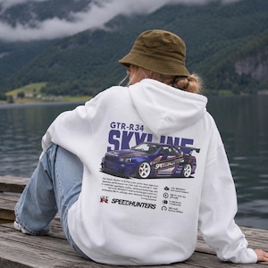 Op de afbeelding: Een wit sweatshirt met capuchon en een grote afbeelding op de achterkant van een blauwe Nissan Skyline GTR-R34 auto. De print bevat de tekst GTR-R34 SKYLINE, technische specificaties en het Speedhunters-logo in zwart, blauw en wit.