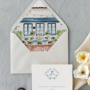 May include: A cream-colored envelope with a watercolor illustration of a courtyard featuring a fountain, blue doors, and string lights on the interior flap. A matching white card displays the monogram A and the text Fiona Tamburini and.
