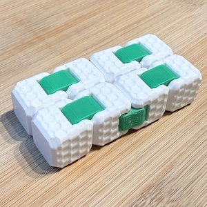 May include: A white and green fidget toy constructed from multiple interlocking rectangular blocks. The white blocks feature a textured, grid-like surface pattern, while the green blocks are smooth and positioned between the white segments.