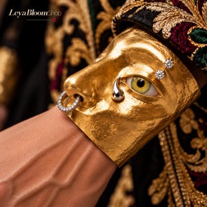 May include: A gold-toned cuff bracelet featuring a sculpted human face with a textured finish. The face includes a realistic eye with a green iris, a silver-toned teardrop accent, two small circular crystals above the eye, and a crystal-encrusted nose ring.