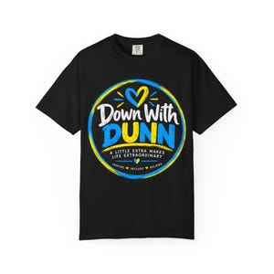 Op de afbeelding: Een zwart t-shirt met korte mouwen en een cirkelvormige afbeelding met blauwe en gele accenten. De tekst luidt Down With DUNN, A LITTLE EXTRA MAKES LIFE EXTRAORDINARY en INSPIRE INCLUDE BELONG, vergezeld door kleine hartjes.