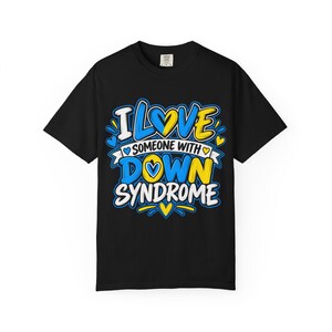 May include: A black short-sleeve t-shirt featuring the text I LOVE SOMEONE WITH DOWN SYNDROME in blue, yellow, and white. The design includes heart icons and decorative flourishes in a bold, graphic style centered on the front of the garment.