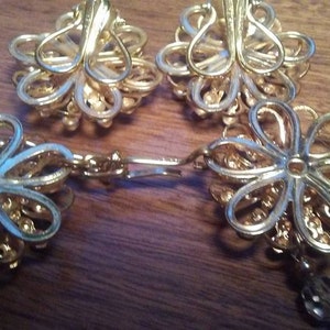 K&ouml;nnte beinhalten: Ein Paar goldfarbene Ohrringe mit Blumenmuster. Die Ohrringe haben eine Kette mit kleinen Perlen, die daran h&auml;ngen.