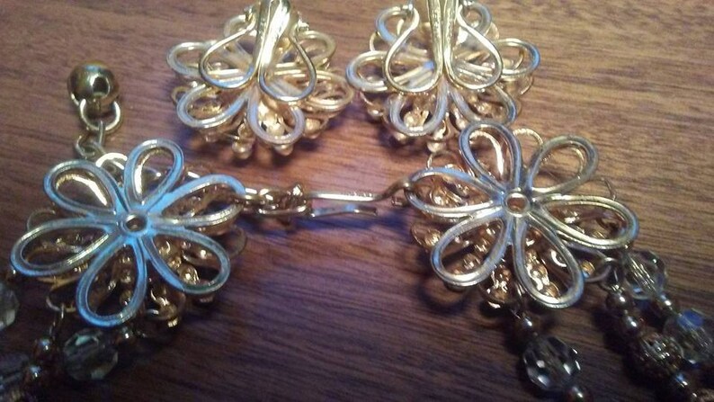 K&ouml;nnte beinhalten: Ein Paar goldfarbene Ohrringe mit Blumenmuster. Die Ohrringe haben eine Kette mit kleinen Perlen, die daran h&auml;ngen.