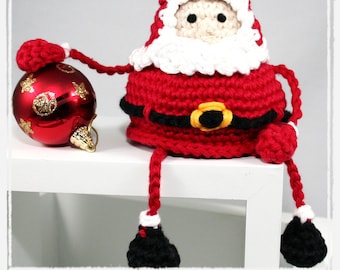 Puede incluir: Una figura de Papá Noel hecha a mano en ganchillo en rojo, blanco y negro, con una hebilla dorada. El Papá Noel está sentado junto a una bola de Navidad roja con diseños de estrellas doradas. El texto "crochet pattern" está en la parte inferior.