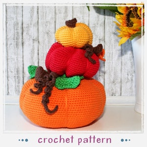 Puede incluir: Una pila de tres calabazas de ganchillo en naranja, rojo y amarillo. Las calabazas tienen tallos marrones y hojas verdes. La calabaza inferior es naranja, la del medio es roja y la superior es amarilla. El texto "crochet pattern" es visible.
