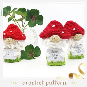Könnte beinhalten: Drei gehäkelte Gnom-Ornamente mit roten Pilzmützen und weißen Bärten. Die Gnome halten Schilder, auf denen "Happy New Year", "Frohes Neues Jahr" und ein Smiley stehen.