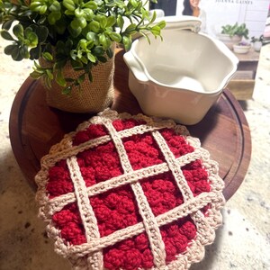 May include: A round, crocheted coaster designed to resemble a cherry pie with a deep red textured center and a light beige lattice crust border. The coaster is placed on a circular wooden board next to a small white ceramic bowl and a green plant.