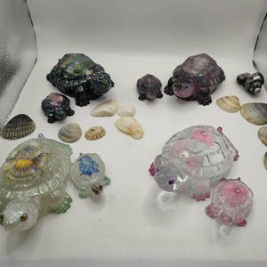 May include: A collection of resin turtle figurines in various sizes and colours, including black, clear, pink, and iridescent finishes, arranged alongside several natural seashells on a white surface. The turtles feature detailed shells.