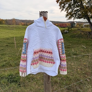 May include: A white quilted hoodie featuring a large front kangaroo pocket and long sleeves with a colorful diamond-patterned patchwork design. The main body of the garment has a subtle floral quilted texture, and the hood has a yellow trim.