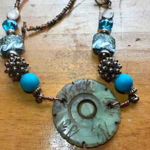 May include: A green jade pendant necklace with a circular design and intricate carvings. The necklace features turquoise and brown beads, as well as silver and copper accents.
