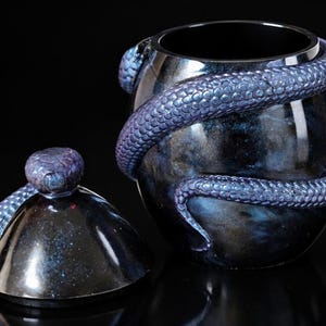 Könnte beinhalten: Ein dunkles, kugelförmiges Gefäß mit abnehmbarem Deckel, um das sich eine texturierte, schillernde blau-violette Schlange windet. Das Gefäß hat eine glänzende, marmorierte Oberfläche in tiefen Blau- und Schwarztönen mit detaillierten Schuppen.