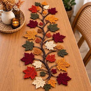 Può includere: Un runner da tavola decorativo caratterizzato da una vite centrale intrecciata marrone scuro con foglie d'acero all'uncinetto nei toni del bordeaux, giallo senape, verde bosco e crema, disposte in un motivo simmetrico lungo il runner.