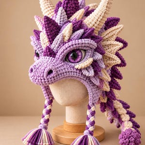 Pode incluir: Um gorro de dragão em croché nos tons roxo, lavanda e creme. O gorro apresenta um padrão de escamas texturizado, dois chifres grandes, uma fila de espigões no topo e duas abas de orelha trançadas que terminam em borlas multicoloridas.