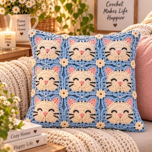 May include: A square crochet pillow cover featuring a repeating pattern of nine cream-colored cat faces with pink ears and black embroidered eyes and whiskers. The cat faces are joined by light blue crochet lattice and small cream-colored flowers.