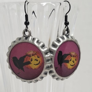 May include: A pair of silver-toned earrings with a black crow and a jack-o'-lantern on a pink background. The earrings are shaped like bottle caps.