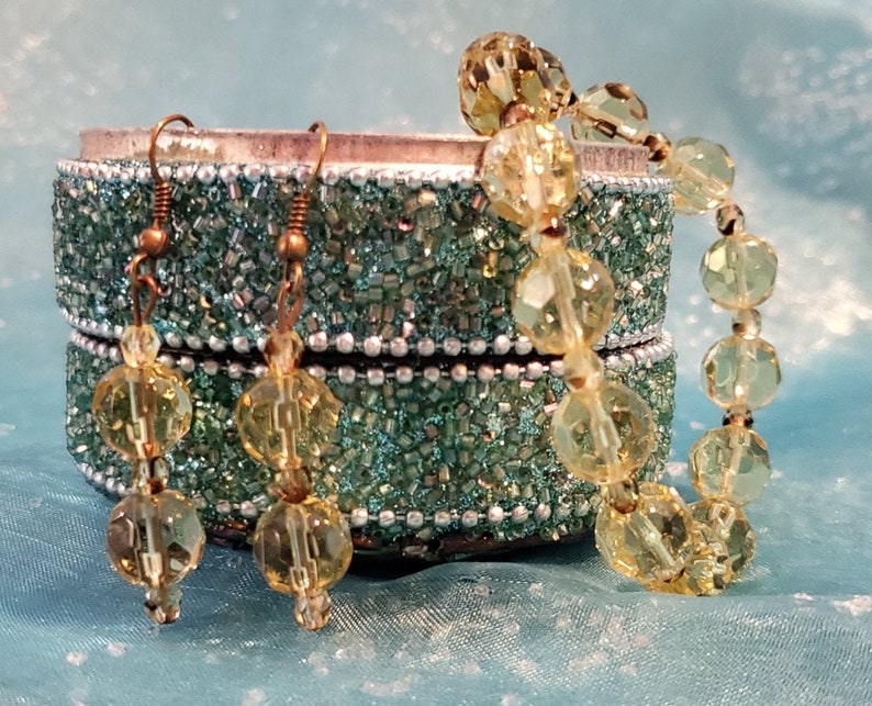 May include: A jewelry set featuring a bracelet and earrings with faceted yellow beads. The earrings have bronze-colored hooks. The bracelet and earrings are displayed next to a small, decorative, turquoise-colored box.