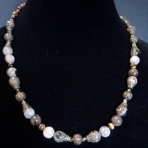May include: A necklace made of brown, white, and gray agate beads with gold and silver accents. The beads are strung on a silver chain.