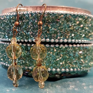May include: A pair of dangle earrings with a vintage aesthetic. Each earring features three faceted, yellow-green beads strung on a copper-colored wire. The earrings are displayed against a teal, beaded jewelry box.
