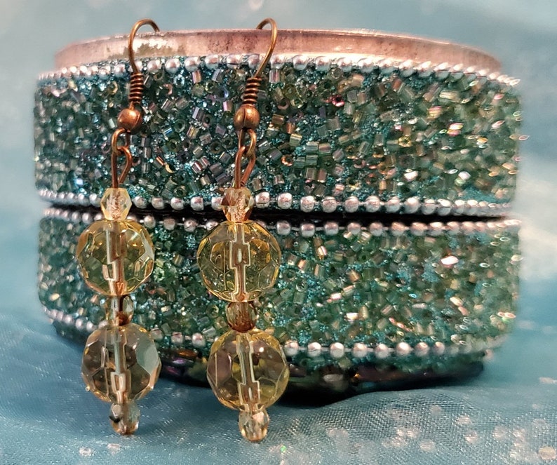 May include: A pair of dangle earrings with a vintage aesthetic. Each earring features three faceted, yellow-green beads strung on a copper-colored wire. The earrings are displayed against a teal, beaded jewelry box.