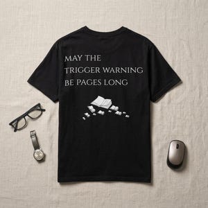 May include: A black short-sleeved t-shirt featuring the white text MAY THE TRIGGER WARNING BE PAGES LONG above a graphic of several open books. The shirt is displayed alongside a pair of black-rimmed glasses, a wristwatch, and a computer mouse.