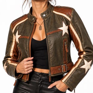 May include: A cropped olive green and brown leather jacket featuring a stand-up collar, silver-tone hardware, and white star appliqués on the chest and sleeves. The jacket includes vertical zippered pockets, a waist belt with a buckle, and light-colored stripes running along the shoulders and down the length of the sleeves.