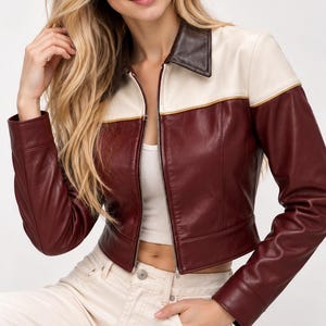 May include: A cropped jacket featuring a dark brown collar, a cream-colored upper chest panel, and a deep burgundy lower body and sleeves. The jacket has a full-length front zipper, long sleeves with cuffs, and a horizontal seam across the chest.