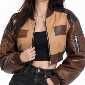 May include: A cropped bomber jacket featuring a color-blocked design in tan, dark brown, and black. The jacket includes a front zipper, ribbed cuffs and collar, a zippered utility pocket on the left sleeve, and rectangular patches on the chest.