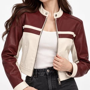May include: A cropped jacket featuring a color-blocked design in deep burgundy and cream. The jacket has a stand-up collar with a snap button, a full-length front zipper, and long sleeves with cream-colored panels along the inner arms.