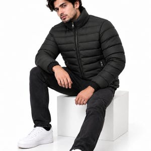 Puede incluir: Una chaqueta acolchada negra con cuello alto, costuras horizontales y cremallera frontal completa. La prenda cuenta con mangas largas con puños acanalados y dos bolsillos laterales con cremalleras plateadas, combinada con pantalones vaqueros negros.