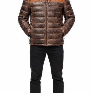 Puede incluir: Una chaqueta acolchada de cuero marrón con cuello alzado, cremallera frontal completa y costuras horizontales. La prenda presenta un canesú de cuero liso color tostado en los hombros y un acabado texturizado y desgastado en la parte inferior.