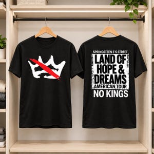 May include: A black t-shirt featuring a white crown graphic with a red diagonal slash on the front. The back displays the text: SPRINGSTEEN E S STREET LAND OF HOPE & DREAMS AMERICAN TOUR NO KINGS in white block lettering on a distressed background.