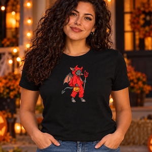 Peut inclure: Un t-shirt noir à manches courtes avec une impression graphique représentant une créature grise vêtue d'un costume de diable rouge avec des ailes, des cornes et une queue, tenant une fourche rouge et un petit seau citrouille orange.