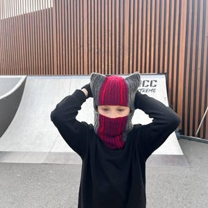 May include: A knitted balaclava featuring cat-like ears on top, designed with vertical stripes in dark grey and deep red. The headgear covers the head and neck, leaving an opening for the eyes, and is worn with a plain black long-sleeved sweatshirt.
