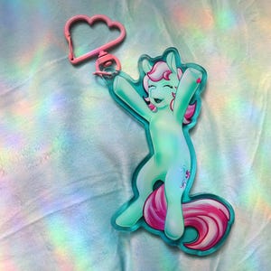 May include: A mint green acrylic keyring featuring a cartoon pony with a pink mane and tail, depicted with arms raised in a joyful pose. The keyring includes a pink cloud-shaped plastic clasp attached to the top of the pony figure.