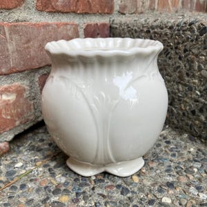 May include: A cream-coloured ceramic planter with a glossy finish, featuring a scalloped rim and embossed leaf-like vertical patterns extending from the base to the upper body. The vessel has a rounded, bulbous shape and a small pedestal base.