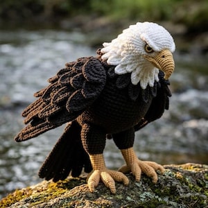 May include: A crocheted bald eagle figurine featuring a white head, a dark brown body with layered wing feathers, and light tan talons. The eagle has a textured, yarn-based construction with a detailed beak and focused eyes, standing upright.