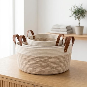 Puede incluir: Un juego de dos cestas de almacenamiento redondas hechas de cuerda enrollada con un diseño de dos tonos, con una sección superior crema y una sección inferior beige. Cada cesta incluye dos asas de piel sintética marrón fijadas con remaches dorados.