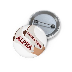 May include: A round pinback button featuring a white background with brown and tan abstract shapes. The text ALPHA is printed in bold red letters with a black outline, and the text LAMBDA THETA is printed in red letters.