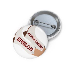 May include: A round pin-back button featuring a white background with a tan graphic element and the red text EPSILON and ALPHA OMEGA. The back of the button is silver-coloured metal with a horizontal pin mechanism for fastening to clothing.