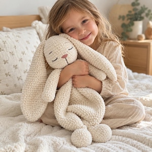 May include: A cream-colored crochet bunny plush toy with long, floppy ears and a soft, textured yarn finish. The bunny features embroidered closed eyes and a small pink stitched nose, creating a simple, neutral appearance for this stuffed animal.