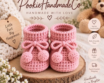 May include: A pair of handmade pink crochet baby booties featuring a ribbed texture, a drawstring tie, and two small pom-poms on the front of each shoe. The booties are displayed on a round wooden slice with text elements visible in the background.