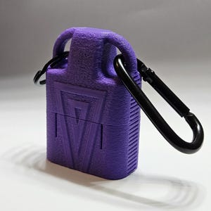 May include: A purple protective case with a textured surface and a geometric triangle design on the front. The case features a top loop attached to a black metal carabiner clip, providing a secure way to carry the rectangular container.