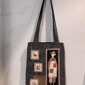 May include: A dark gray canvas tote bag featuring a vertical rectangular patch of a person wearing a hat, plaid shirt, and apron. Additional smaller square patches depict a goat, two glass bottles, and a globe. The bag has two long shoulder straps.