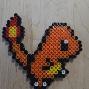 May include: A pixel art figure crafted from orange, black, yellow, and white plastic fuse beads. The design depicts a stylized creature with a large head, a curved tail ending in a flame-like pattern, and small white accents on the feet.
