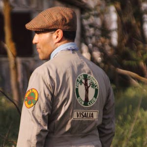 Könnte beinhalten: Ein Mann trägt eine braune und weiße karierte Mütze und eine beige Jacke mit einem Tulare County Gun Club Patch. Der Patch zeigt einen grünen Baum und den Text "Tulare County Gun Club Visalia".